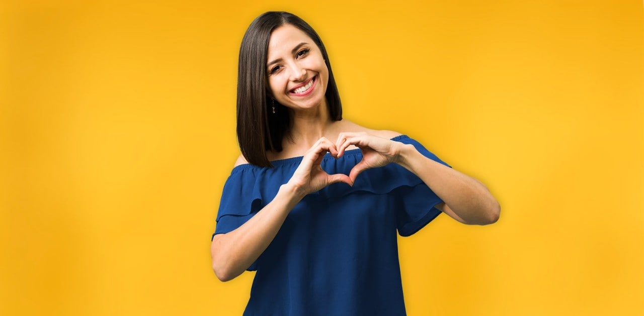 Hispanic woman standing in front of yellow wall with hands forming a heart