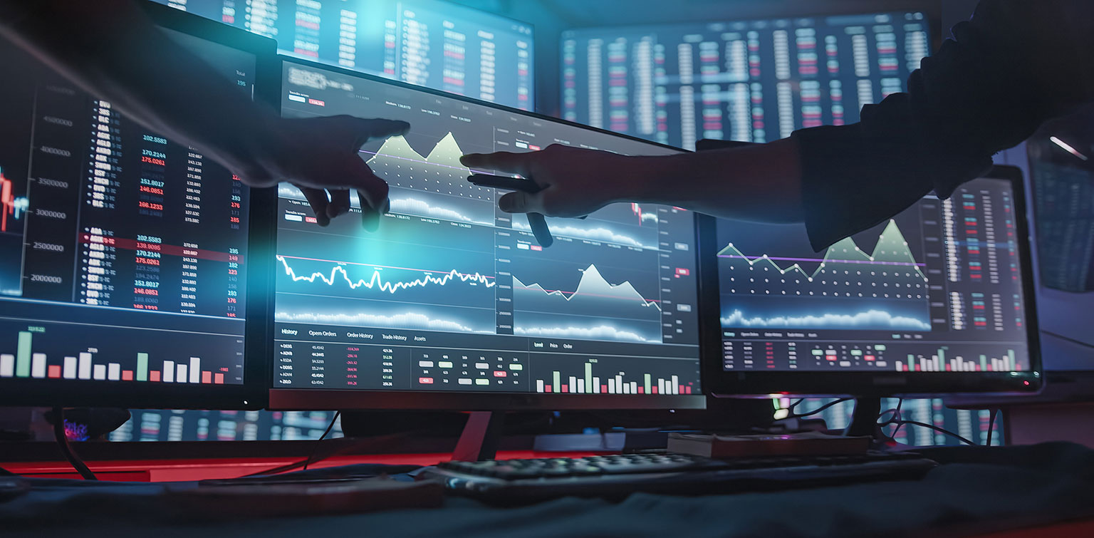 Two analysts pointing to digital monitors displaying financial data