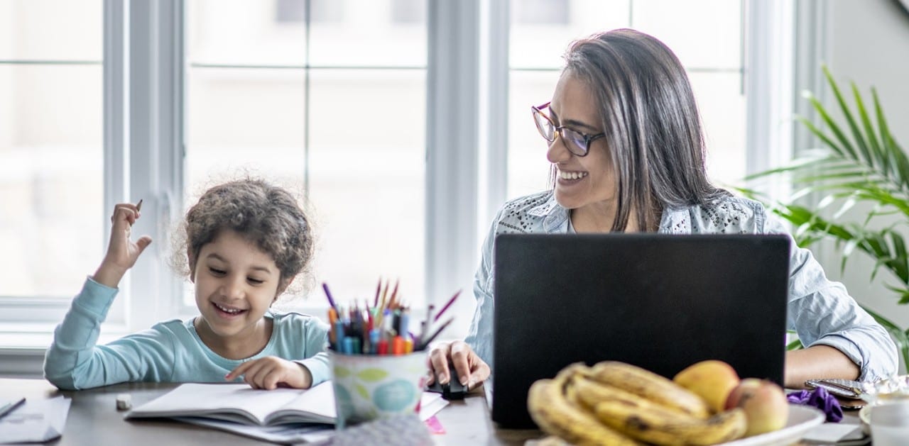 Mother working on laptop sitting next to daughter doing schoolwork
