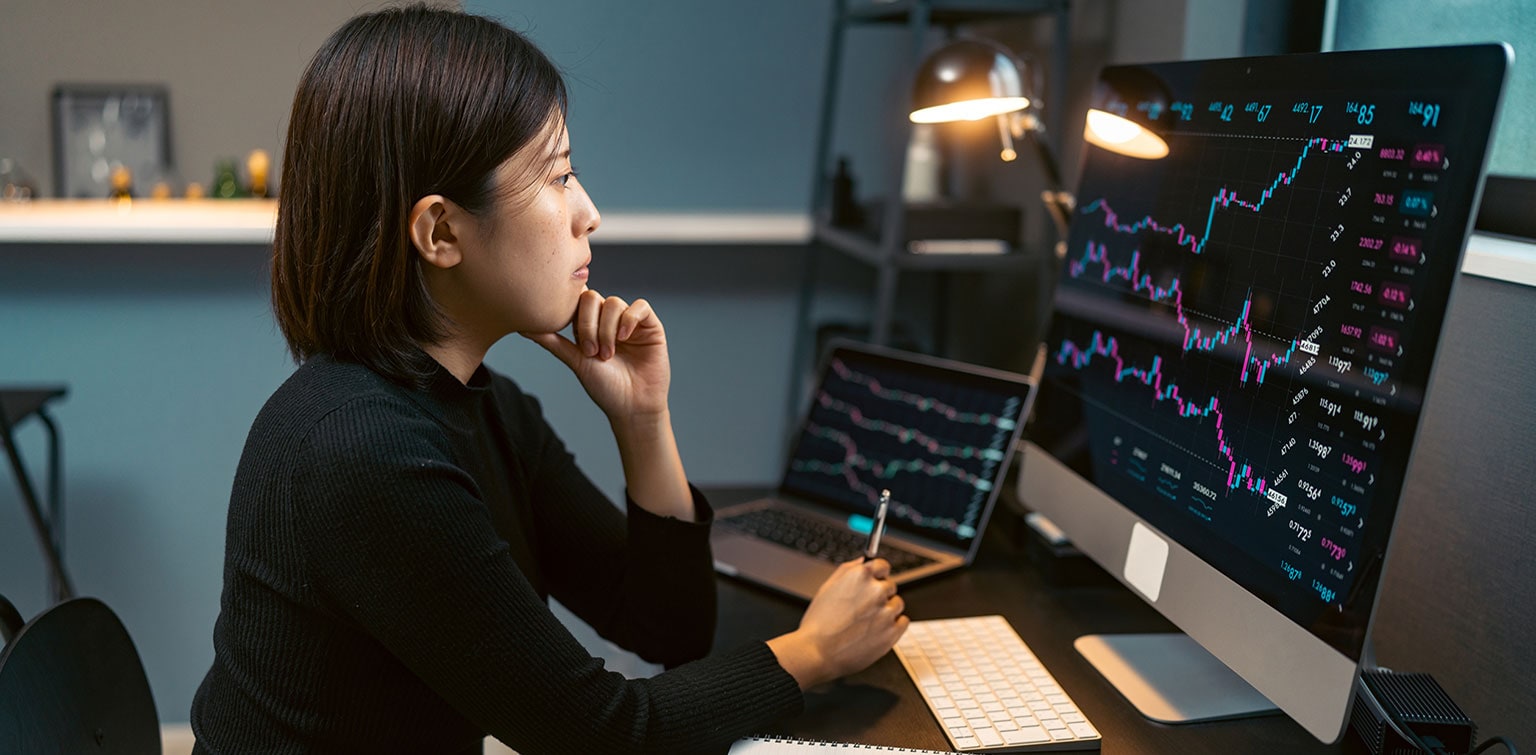 Asian-American female analyst reviewing stock market data on multiple monitors