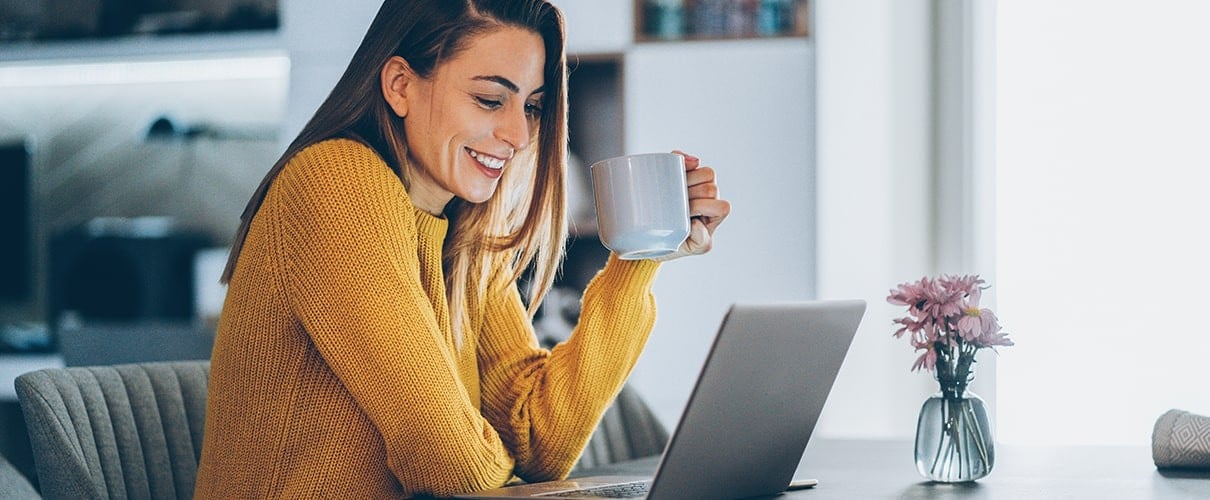 Woman sitting at desk holding coffee mug and looking at laptop