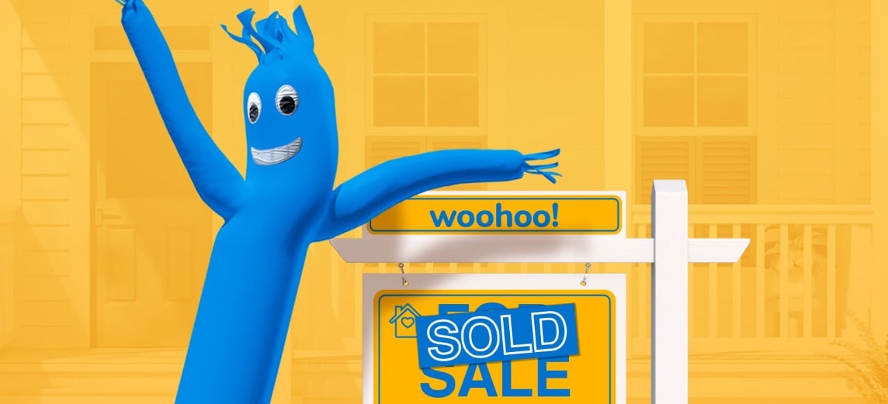 Smiling blue inflatable in front of a "sold" real estate sign on yellow background